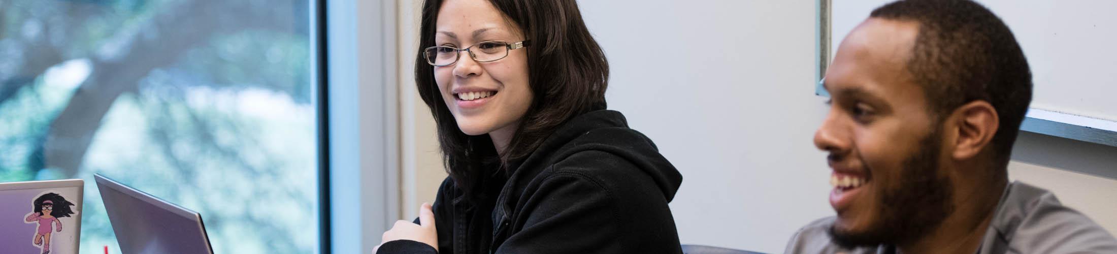 A male and female student smiling looking at their laptops in class.